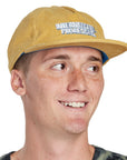 Headshot of a young man wearing a mustard color corduroy hat with the words International Pro Designs printed on the front of the hat in white block letters.