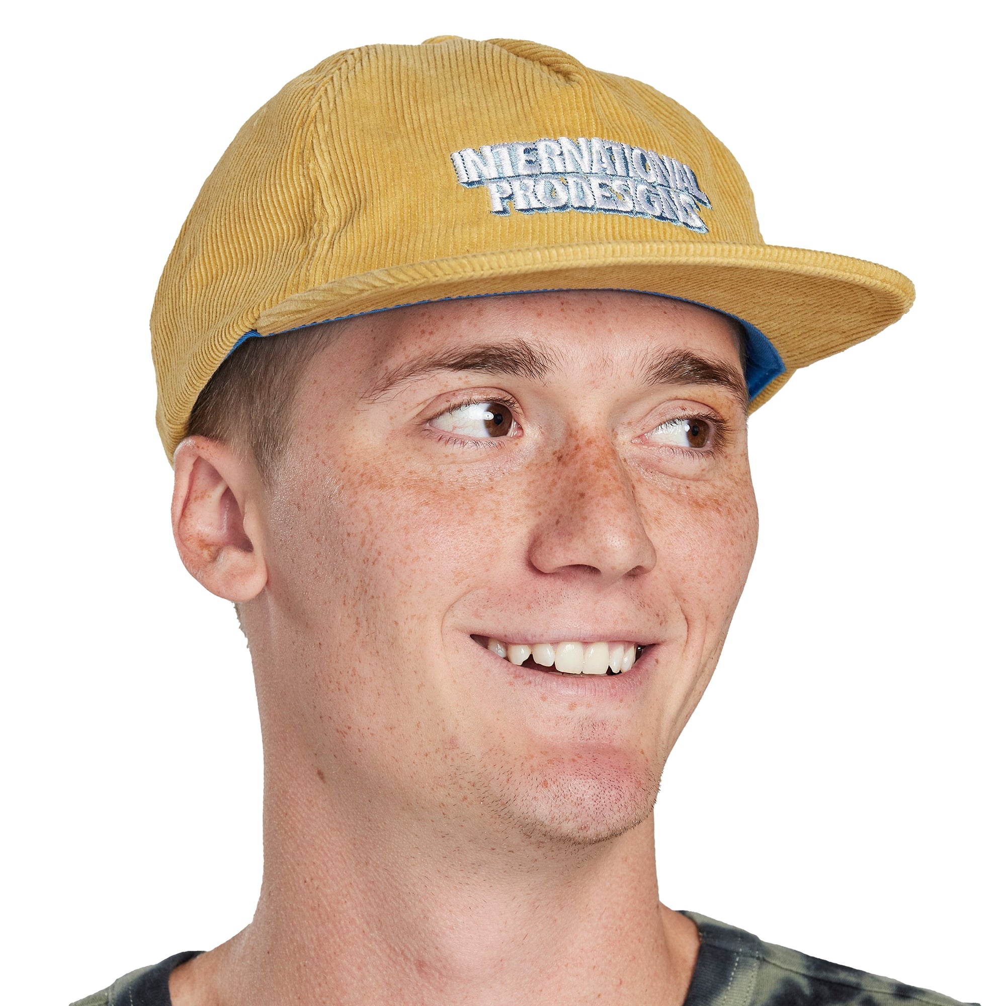 Headshot of a young man wearing a mustard color corduroy hat with the words International Pro Designs printed on the front of the hat in white block letters.