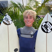 wyatt standing between a couple surfboards wearing an ipd wetsuit with a huge smile on his face