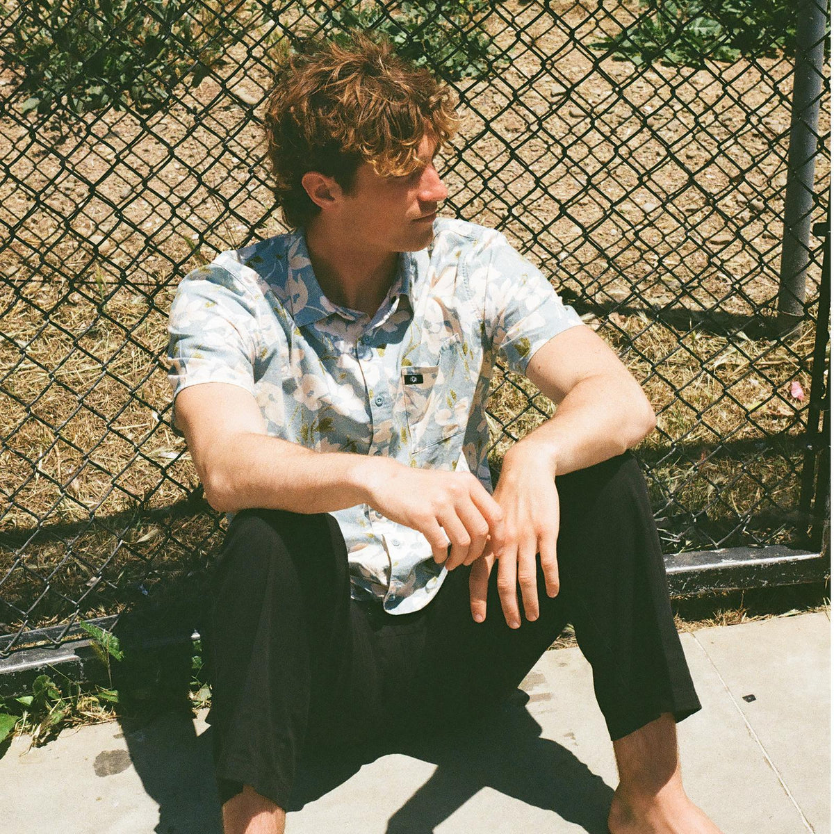 A man wearing a light blue short sleeve floral button-down shirt, sitting relaxed against a chain-link fence. The shirt features a vibrant floral pattern in shades of white and pink. The man&#39;s posture suggests comfort and relaxation as he leans casually against the fence. 