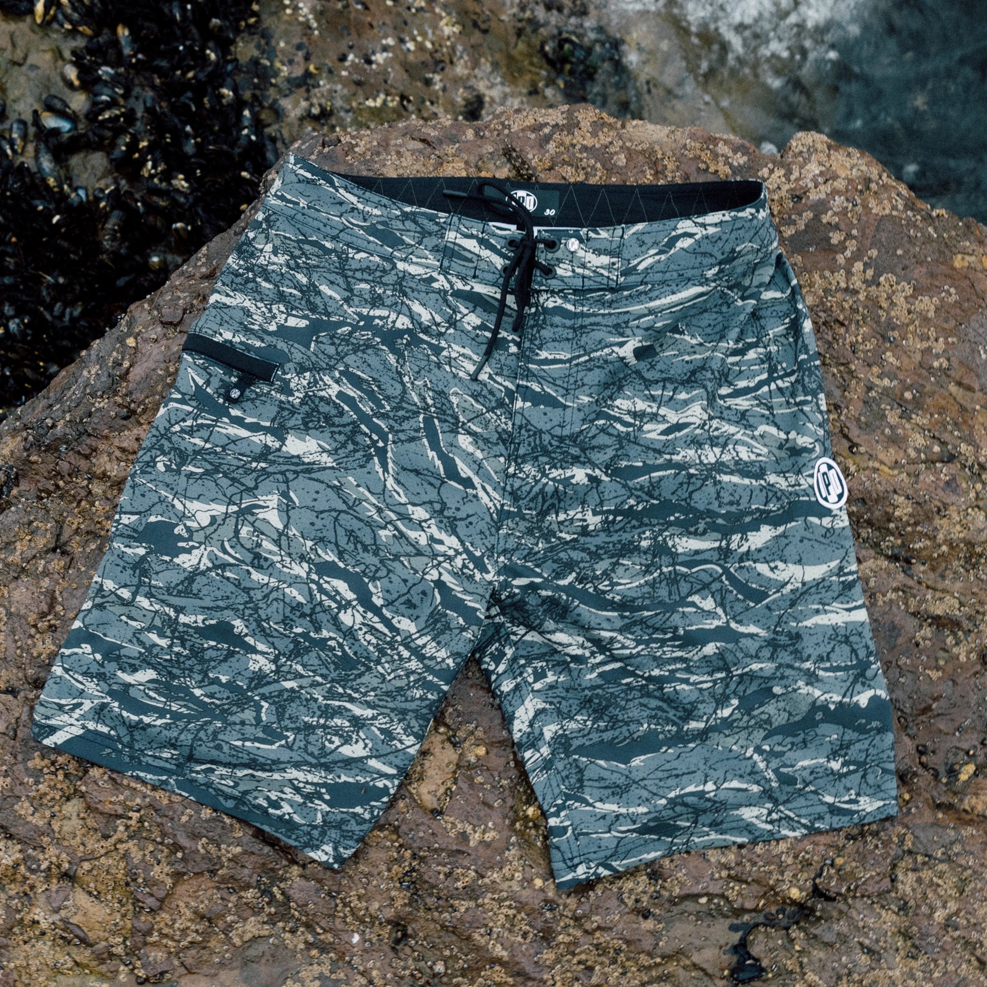 Camouflage board shorts laid out on rocks by the ocean. Made from 86% recycled polyester and 14% spandex. Features a 20-inch straight hem outseam and stretch mesh pocket bag.