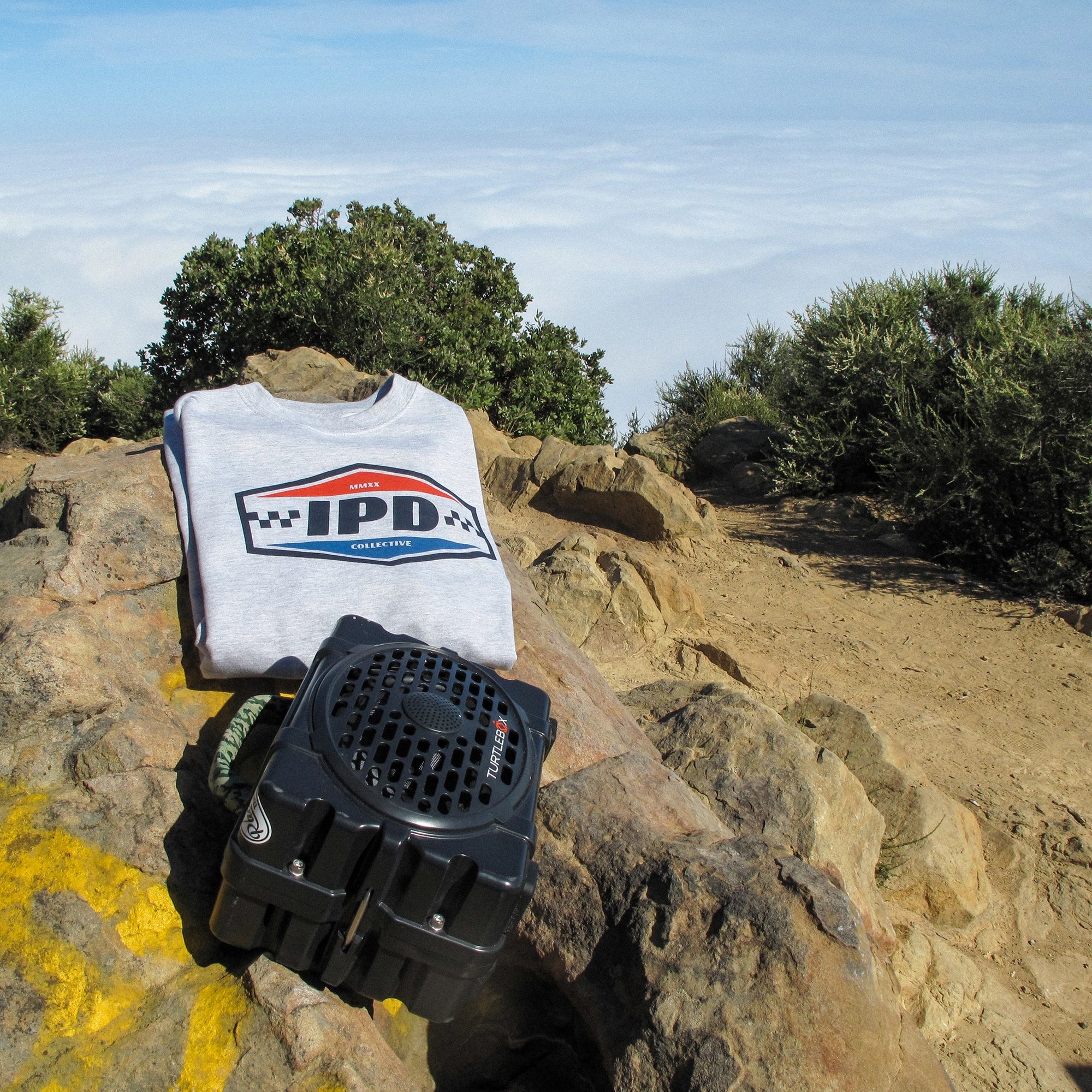 IPD logo sweatshirt and Turtlebox speaker displayed on rocky terrain with a scenic view.