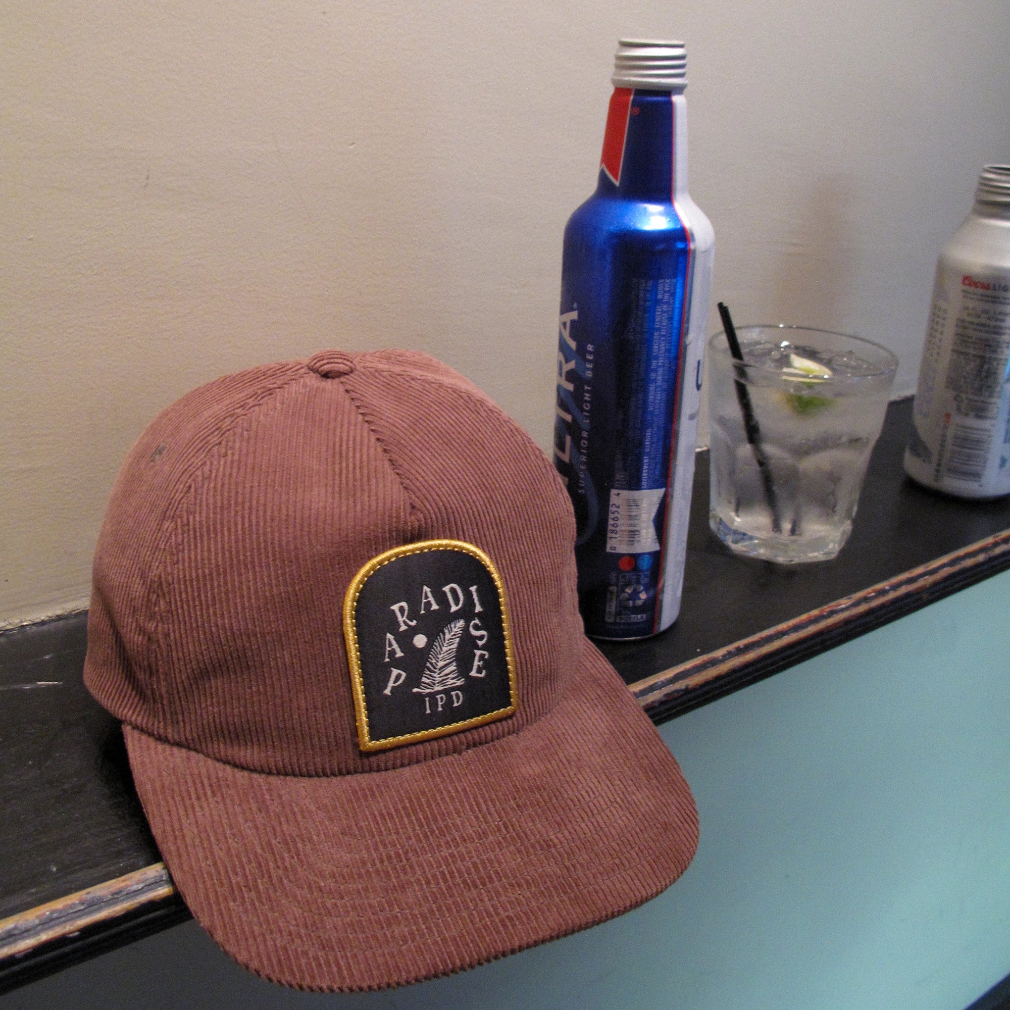 IPD Paradise corduroy hat displayed on a shelf with a drink and beer can in the background