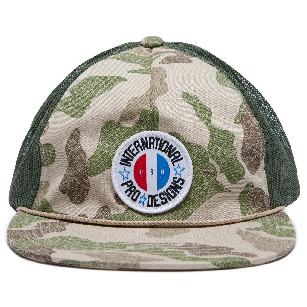 front view of flat brim brigade trucker snapback hat in camo. Showing front logo of international pro designs surrounding circular USA logo 