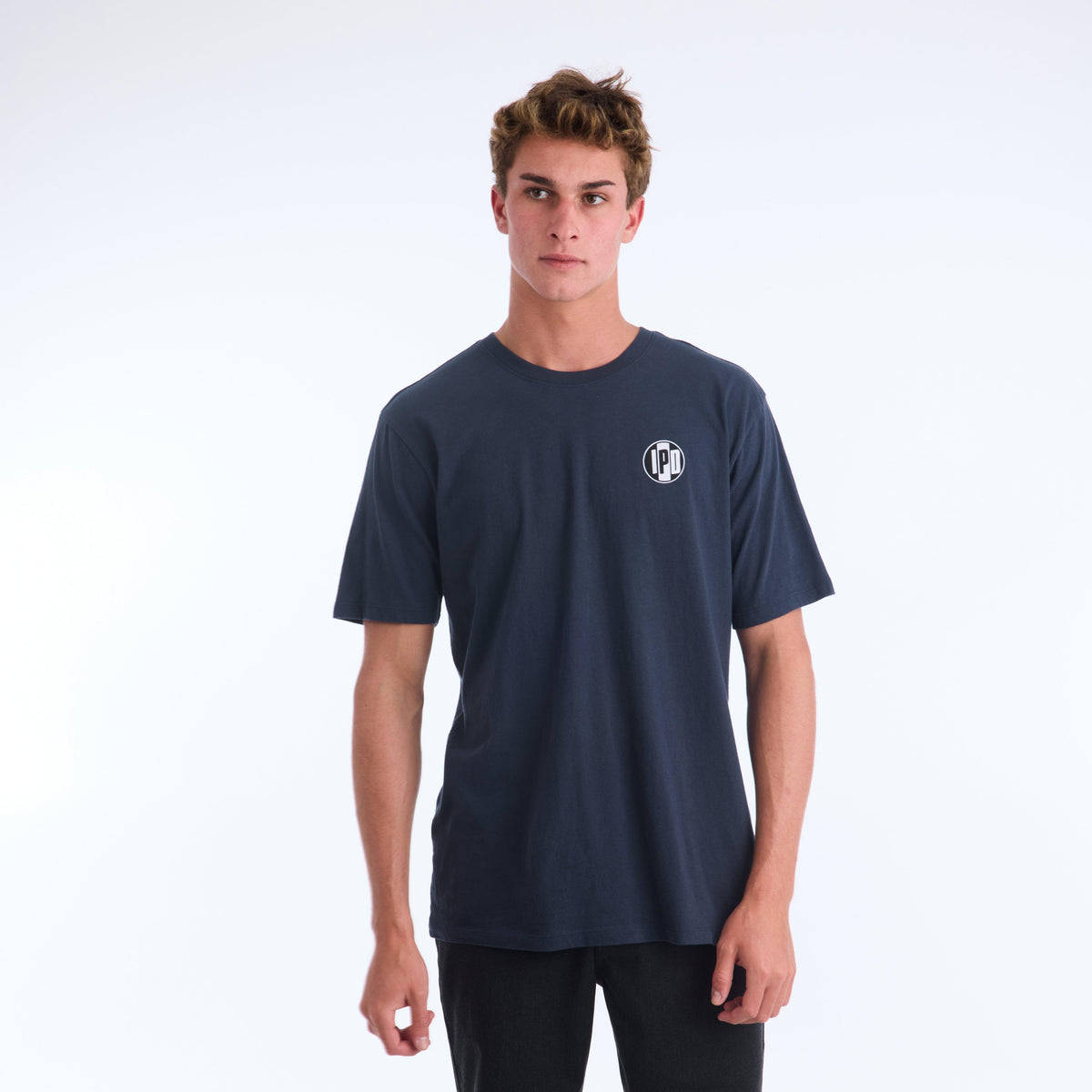 The Surf Shop Super Soft Tee has a left chest front graphic of the IPD logo in black and light blue. The back pictures a hand-drawn depiction of a surf shop across the upper middle back of the shirt. The body color of the shirt is navy and the graphic color is multicolored and has a small black side seam label with the IPD logo.