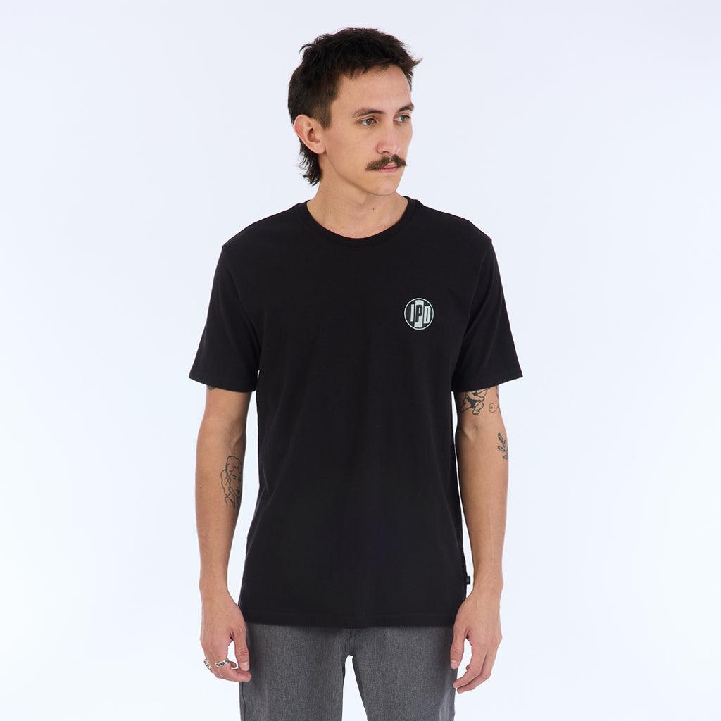 The Surf Shop Super Soft Tee has a left chest front graphic of the IPD logo in black and light blue. The back pictures a hand-drawn depiction of a surf shop across the upper middle back of the shirt. The body color of the shirt is black and the graphic color is multicolored and has a small black side seam label with the IPD logo.