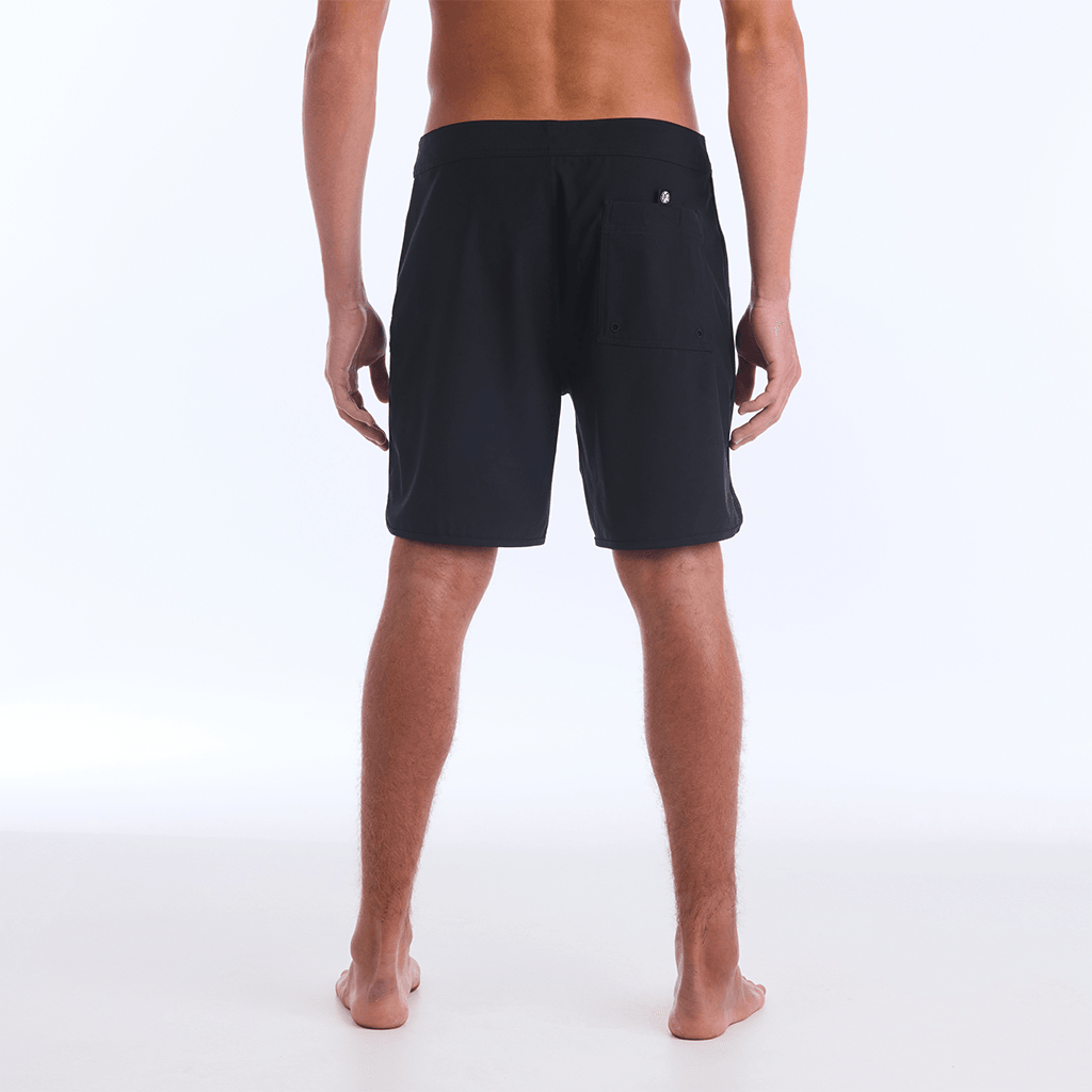 The HI 83 Solid Scallop boardshort on male model features an 18” length and an all black coloring, delivering maximum comfort in a minimalist look. It also has a waistband tie, back patch pocket, and a black IPD logo patch halfway up the left leg taping