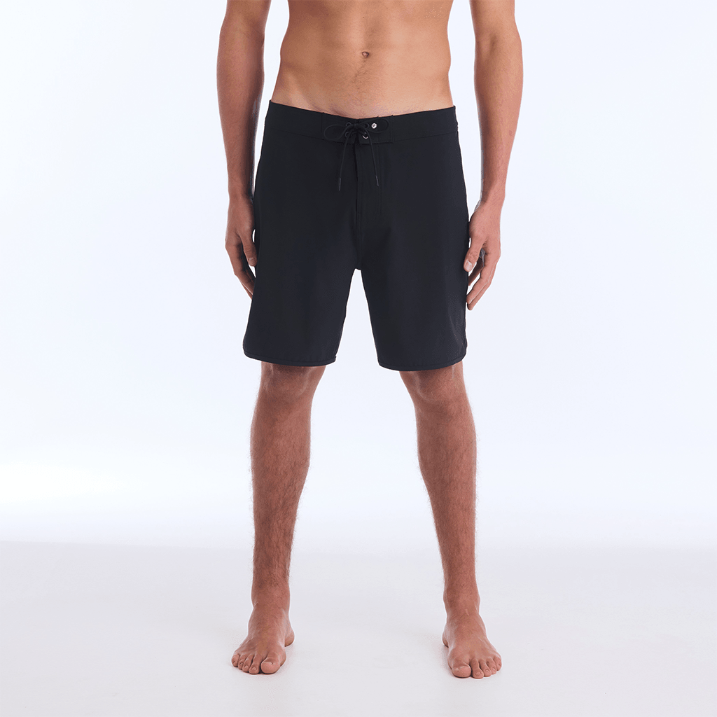 The HI 83 Solid Scallop boardshort on male model features an 18” length and an all black coloring, delivering maximum comfort in a minimalist look. It also has a waistband tie, back patch pocket, and a black IPD logo patch halfway up the left leg taping