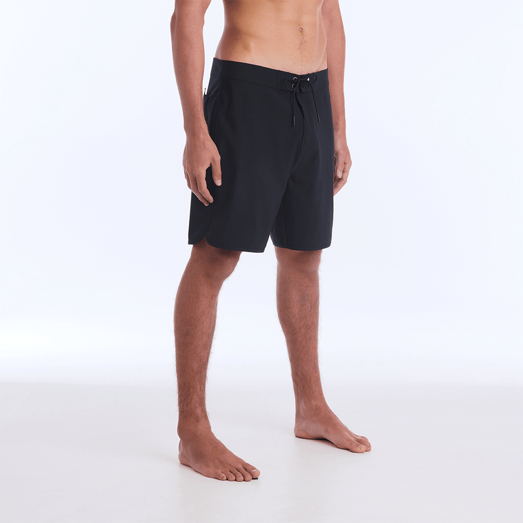 The HI 83 Solid Scallop boardshort on male model features an 18” length and an all black coloring, delivering maximum comfort in a minimalist look. It also has a waistband tie, back patch pocket, and a black IPD logo patch halfway up the left leg taping